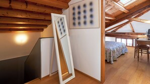 A room with a sloped ceiling, wooden beams, a bed with a floral bedspread, a desk with a chair, and a framed picture on the wall. - Ca' dell'Arte - San Giacomo (Venezia)