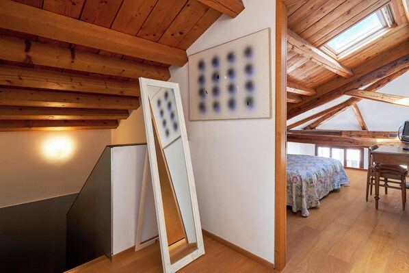 Una habitación con techo inclinado, vigas de madera, una cama con colcha estampada, un escritorio con silla y un cuadro en la pared.