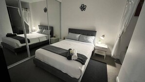 Ein modernes Hotelzimmer mit einem großen Bett, einem Nachttisch mit Lampe, einem Spiegel und einer kleinen Topfpflanze.