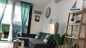 Ein modernes Wohnzimmer mit einem grauen Sofa, einem kleinen weißen Beistelltisch, einer Stehlampe und einem hölzernen Bücherregal.