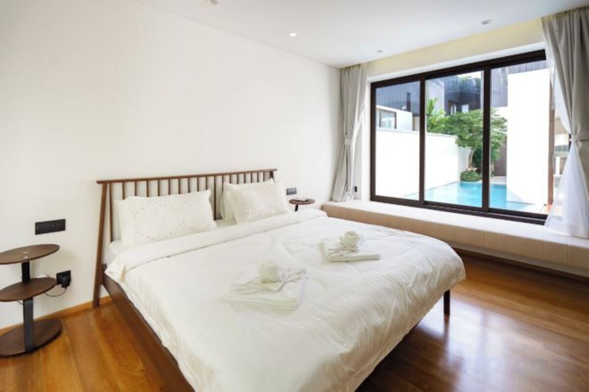 A hotel room with a large bed, a bedside table, a window with a view of a pool, and a small round table.