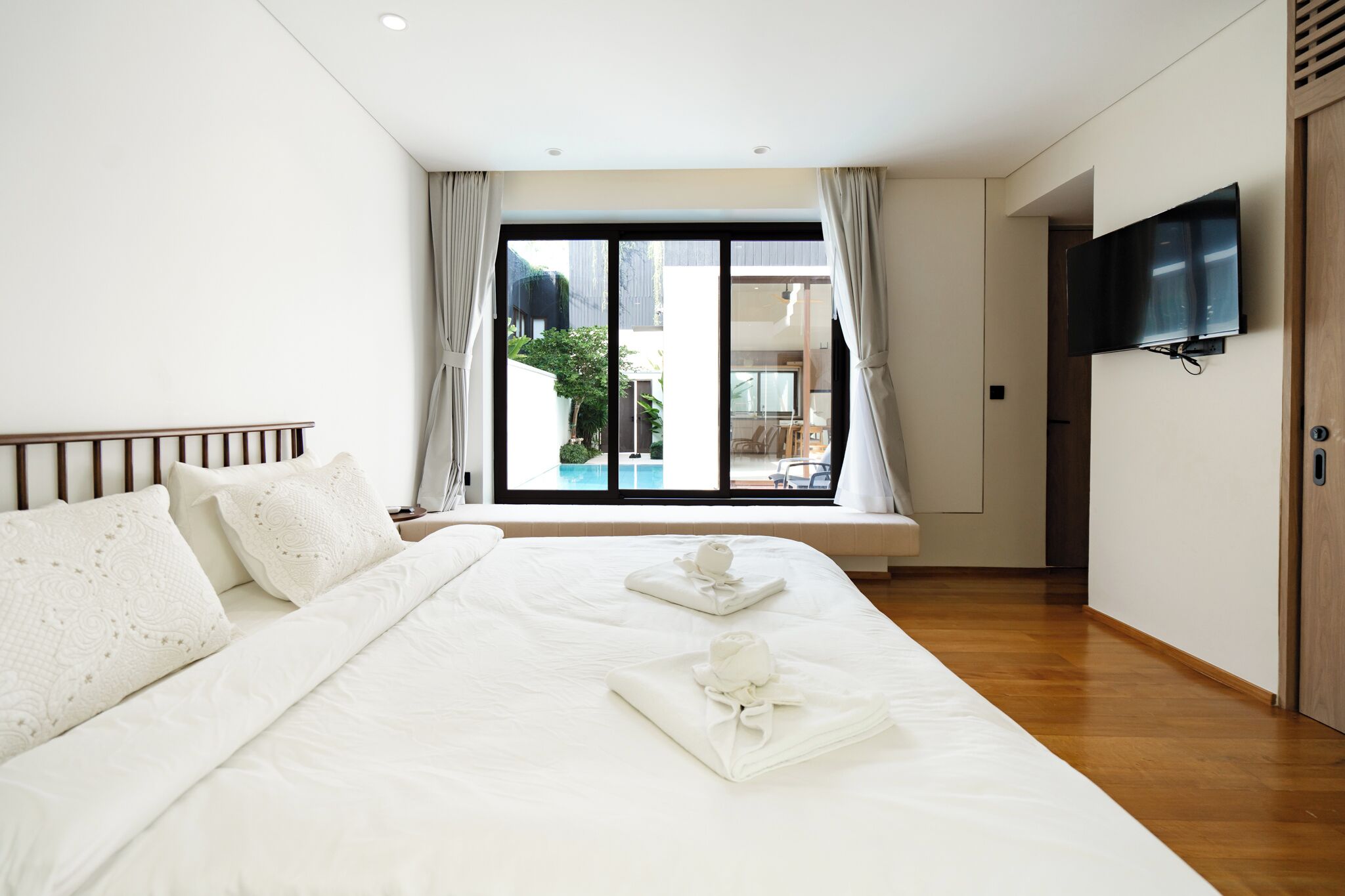 A hotel room with a large bed, a TV mounted on the wall, and a view of a swimming pool through the window.