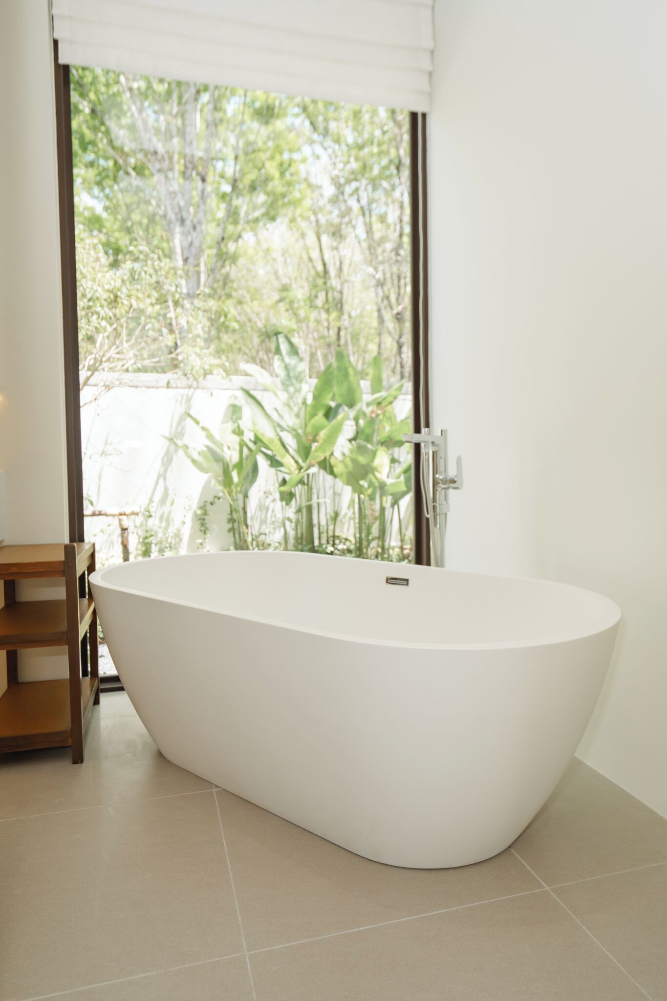 A modern bathroom with a freestanding bathtub, a wooden chair, and a view of greenery outside.