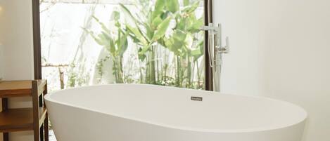 A modern bathroom with a freestanding bathtub, a wooden chair, and a view of greenery outside.