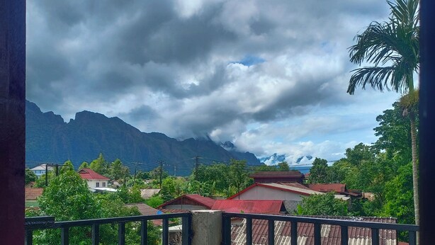 A balcony with a metal railing overlooking a mountainous landscape with houses and greenery. - Dok Champa Hotel (Vang Vieng)