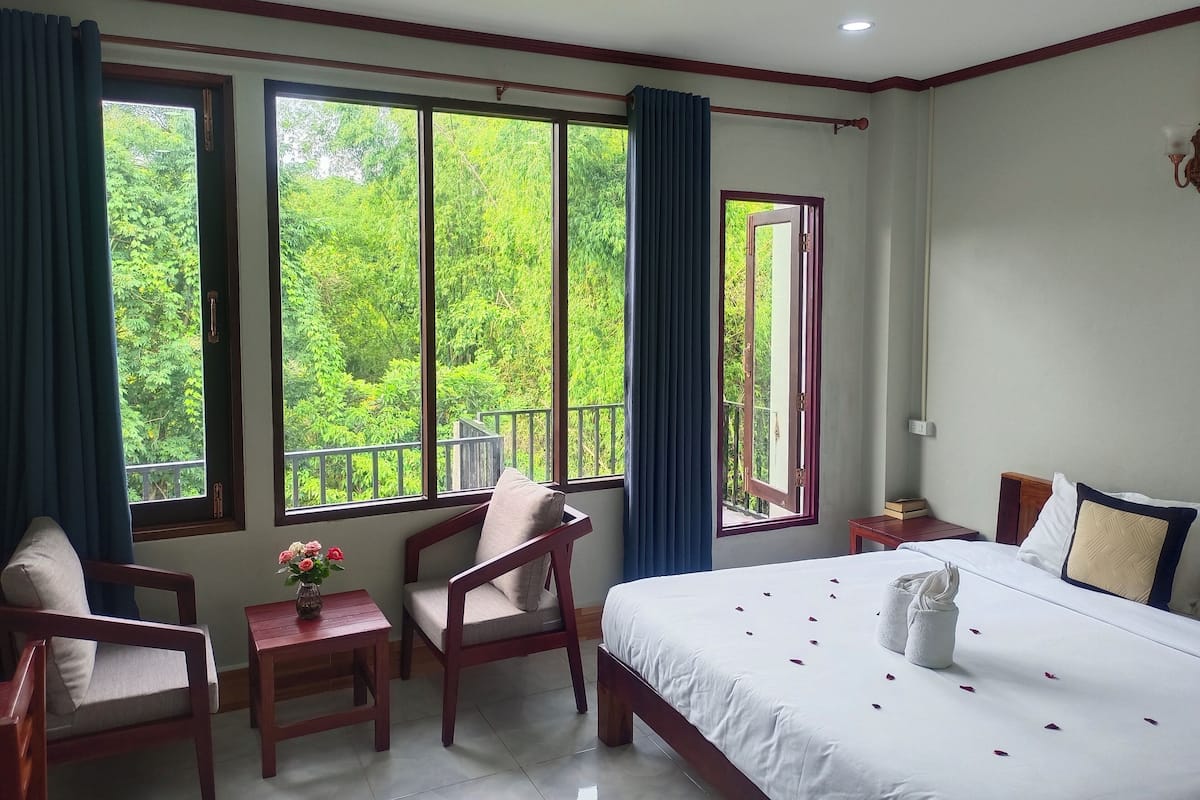 A hotel room with a bed, two chairs, a small table with a vase of flowers, and large windows offering a view of greenery.
