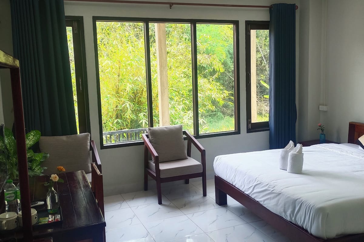 A hotel room with a bed, a chair, a desk, and large windows offering a view of greenery.