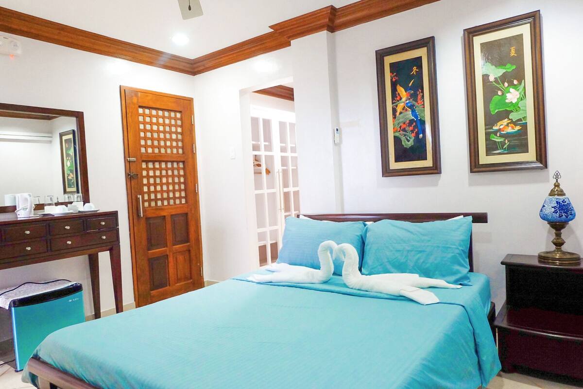 Une chambre d’hôtel comprenant un lit, une tête de lit en bois, un bureau avec une chaise, une armoire et deux œuvres d’art encadrées accrochées au mur.