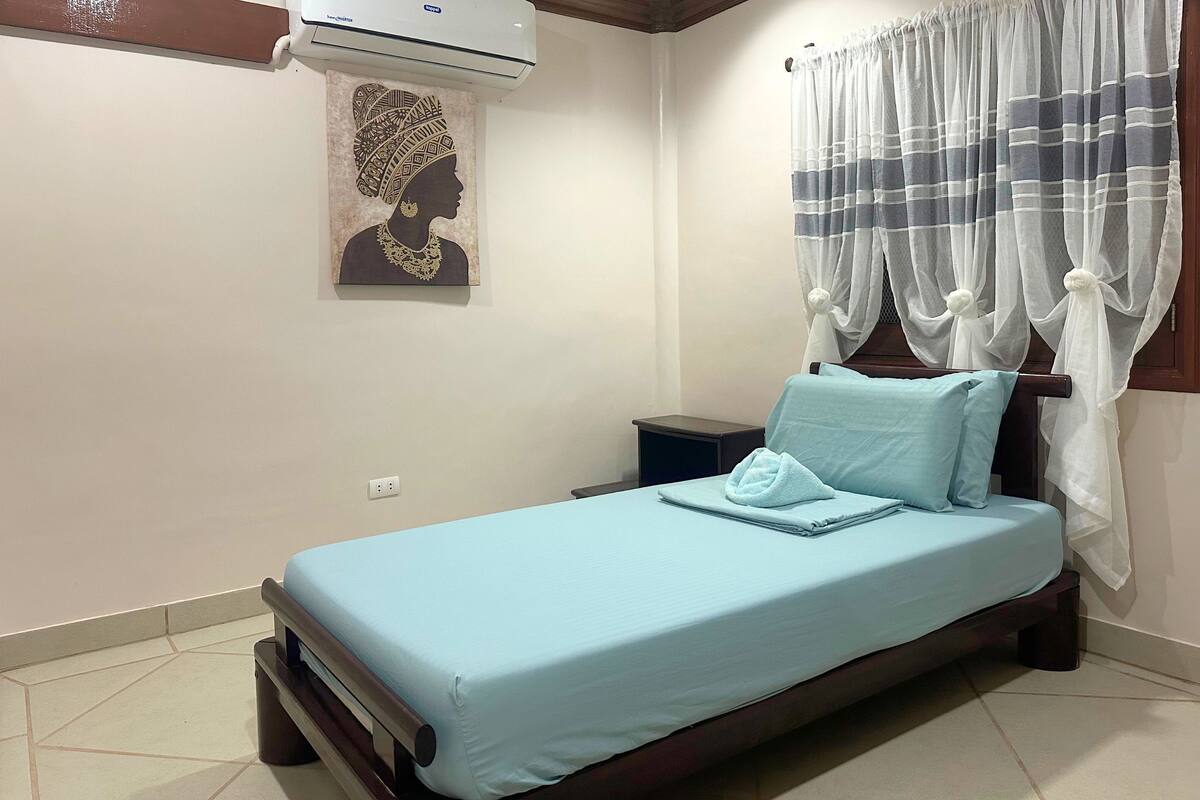 Une chambre à coucher comprenant un lit, une table de chevet, un climatiseur fixé au mur, un rideau et un tableau encadré.