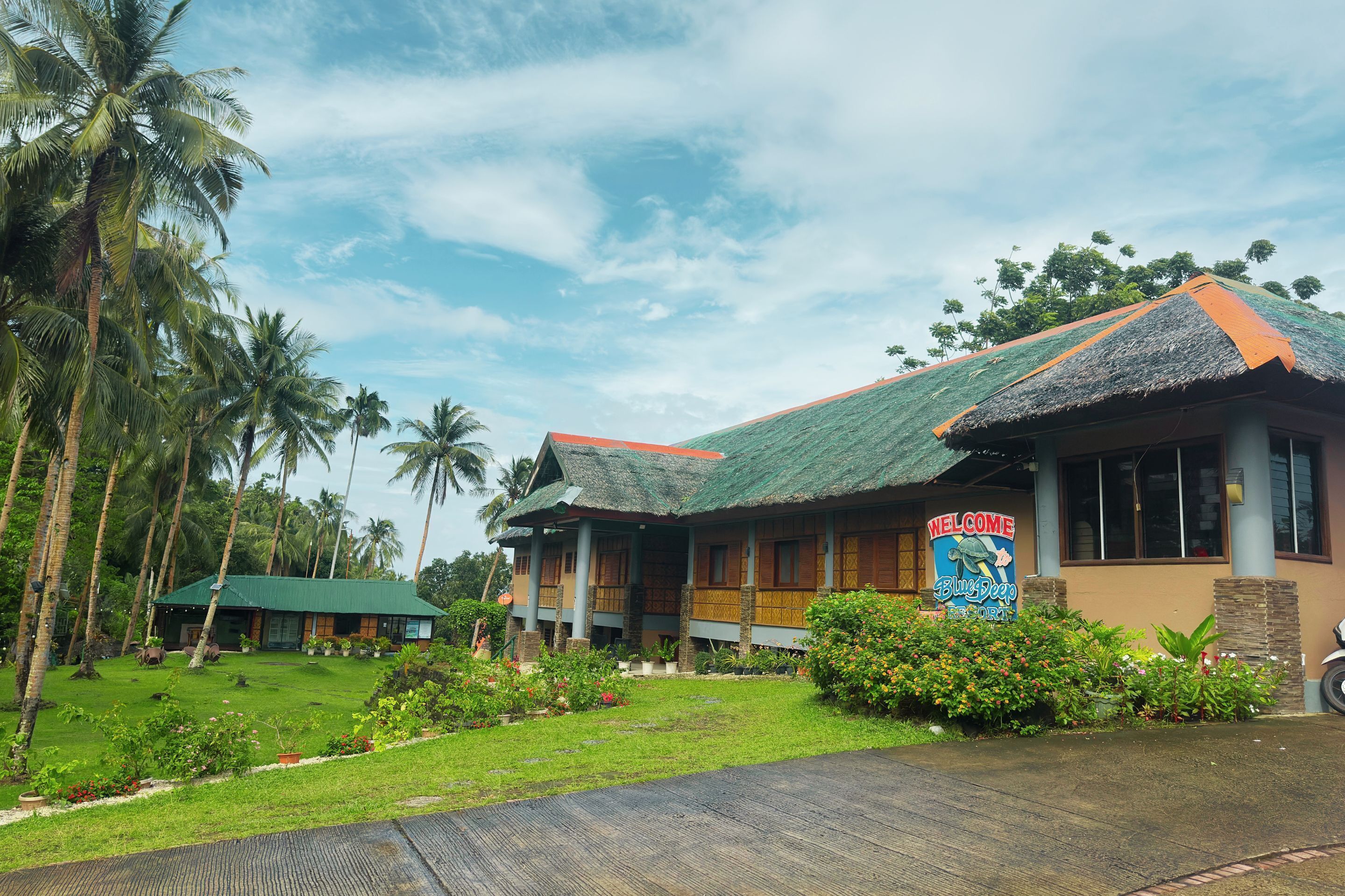 A resort with a thatched roof, a sign reading "Welcome Blue Lagoon", and a well-maintained garden with palm trees and tropical plants.