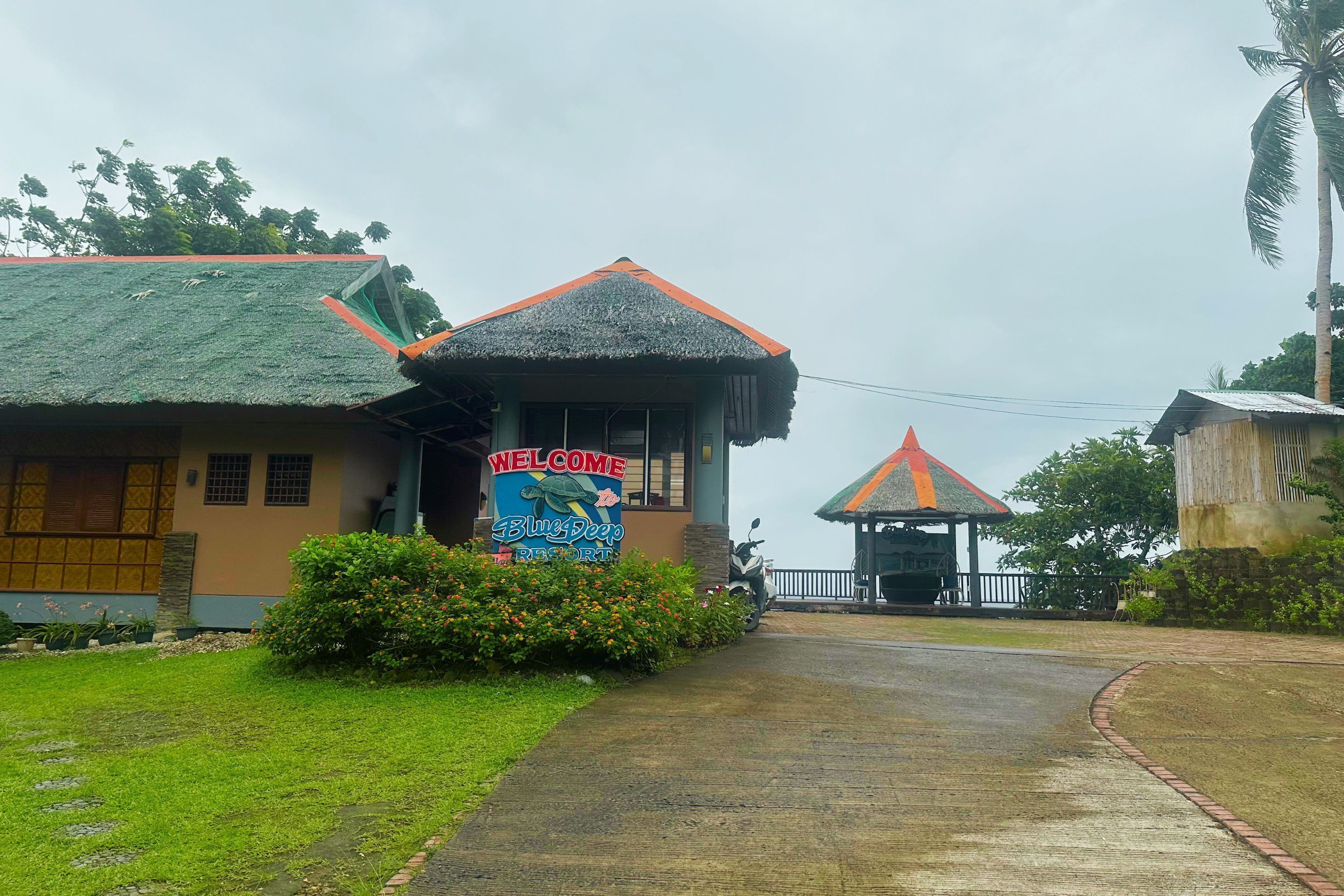 A building with a 'Welcome' sign, a thatched roof, and a pathway leading to a covered entrance.