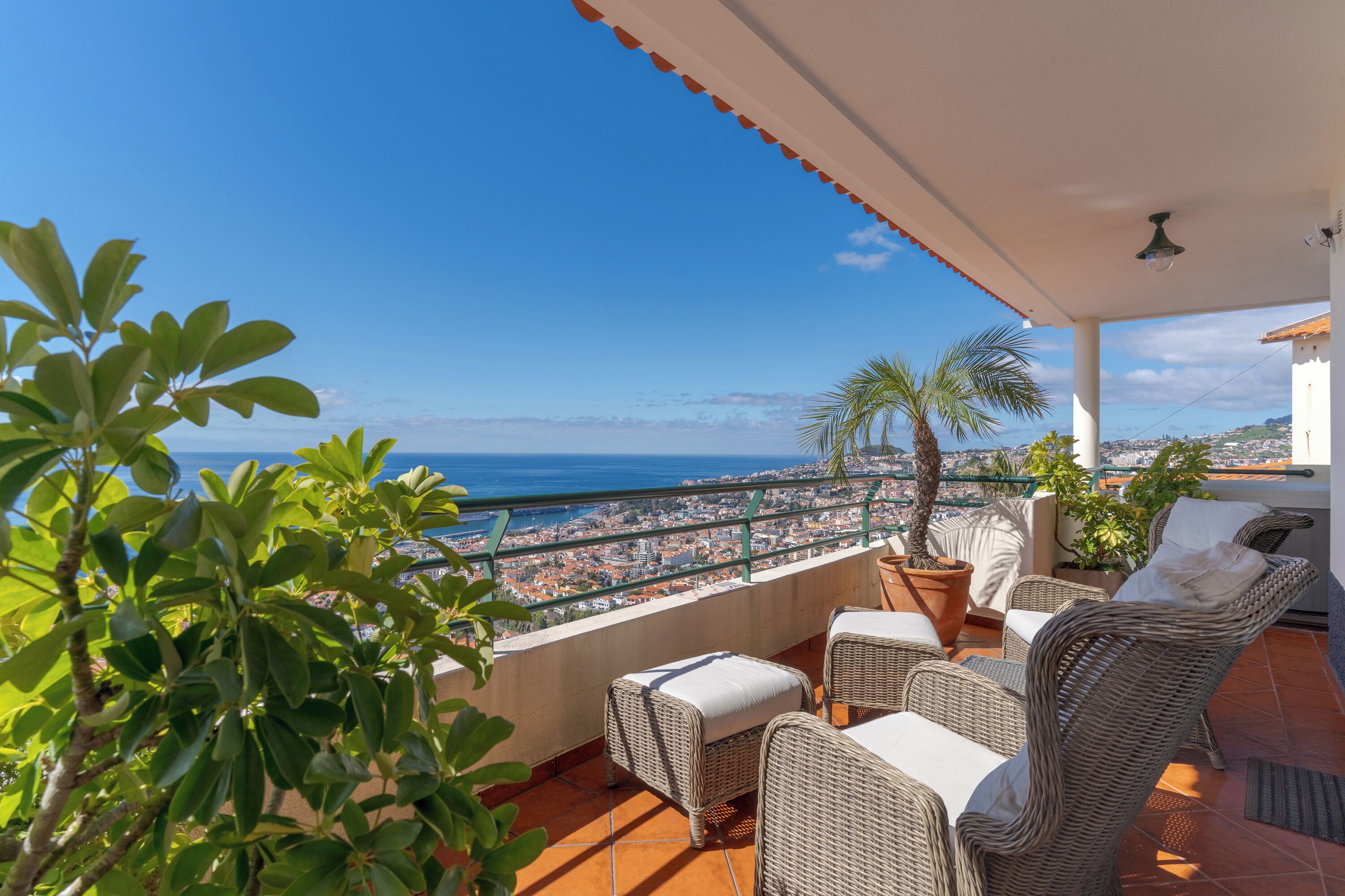 Varanda com móveis de vime, plantas em vasos e vista para a cidade e o mar.