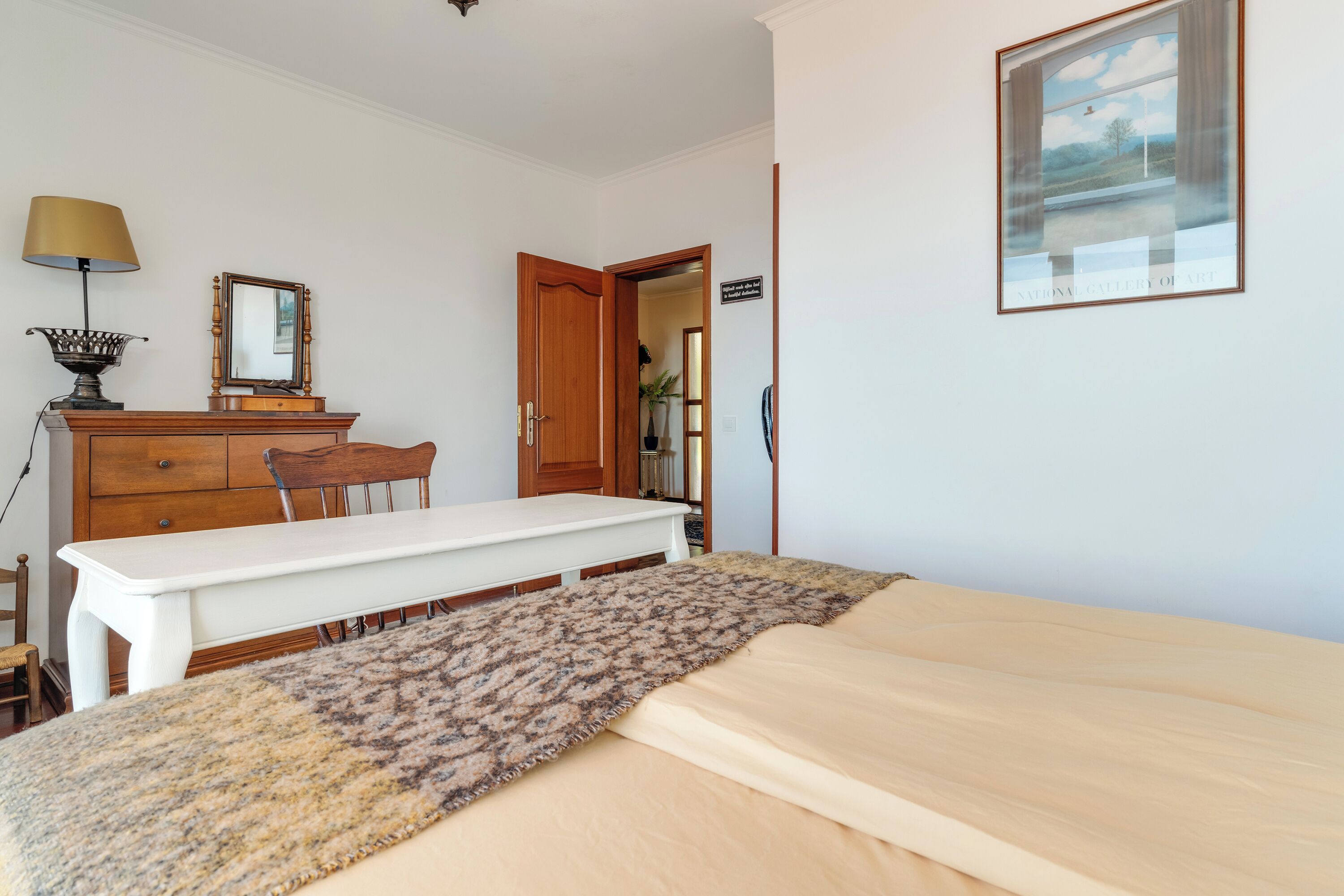 A bedroom with a bed, a desk, a chair, a dresser, a lamp, a mirror, and a framed picture on the wall.