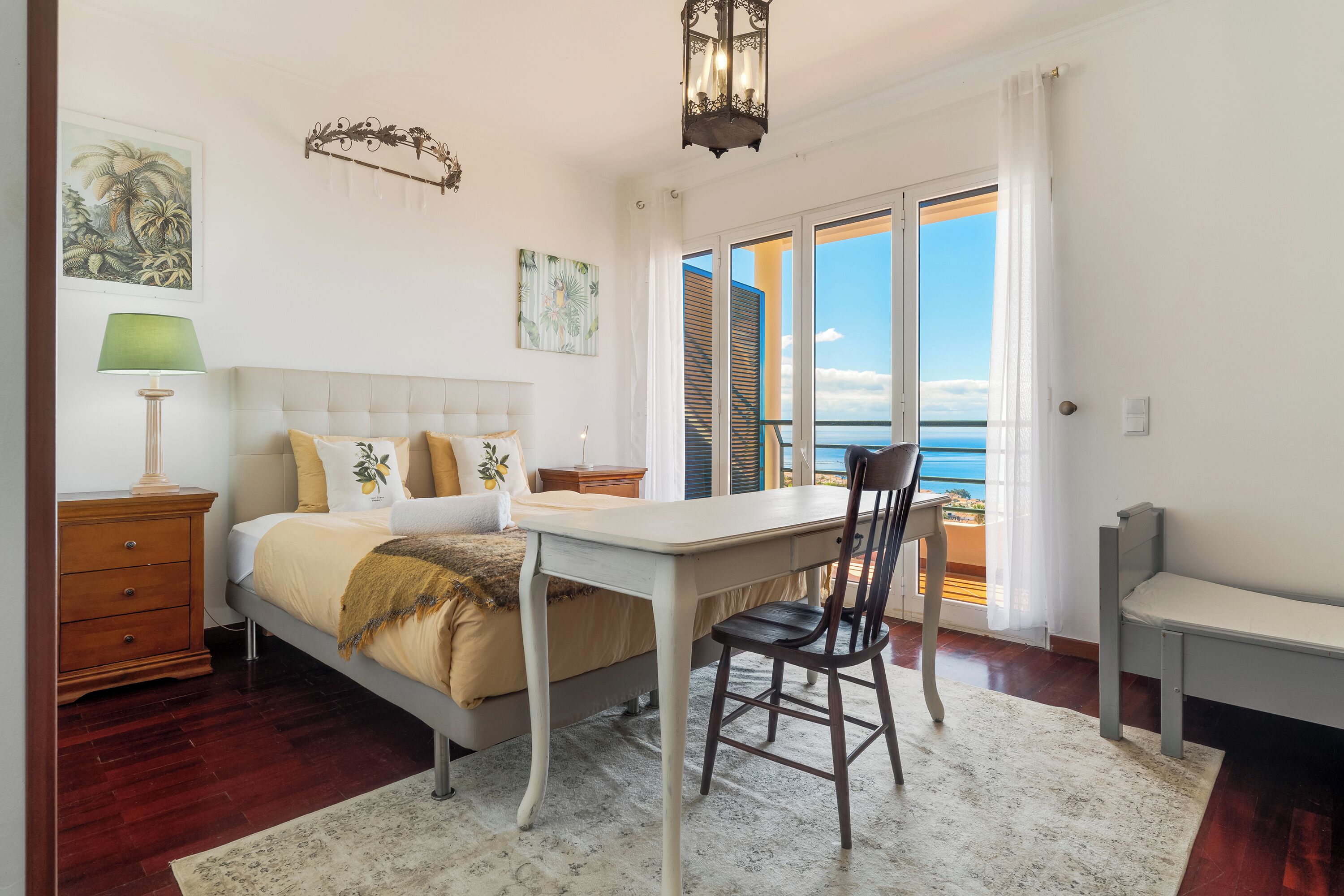 Quarto com cama, escrivaninha, cadeira e vista para o mar através de uma janela grande.