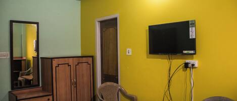 A room with a wooden wardrobe, a mirror, a television mounted on the wall, and two wicker chairs.