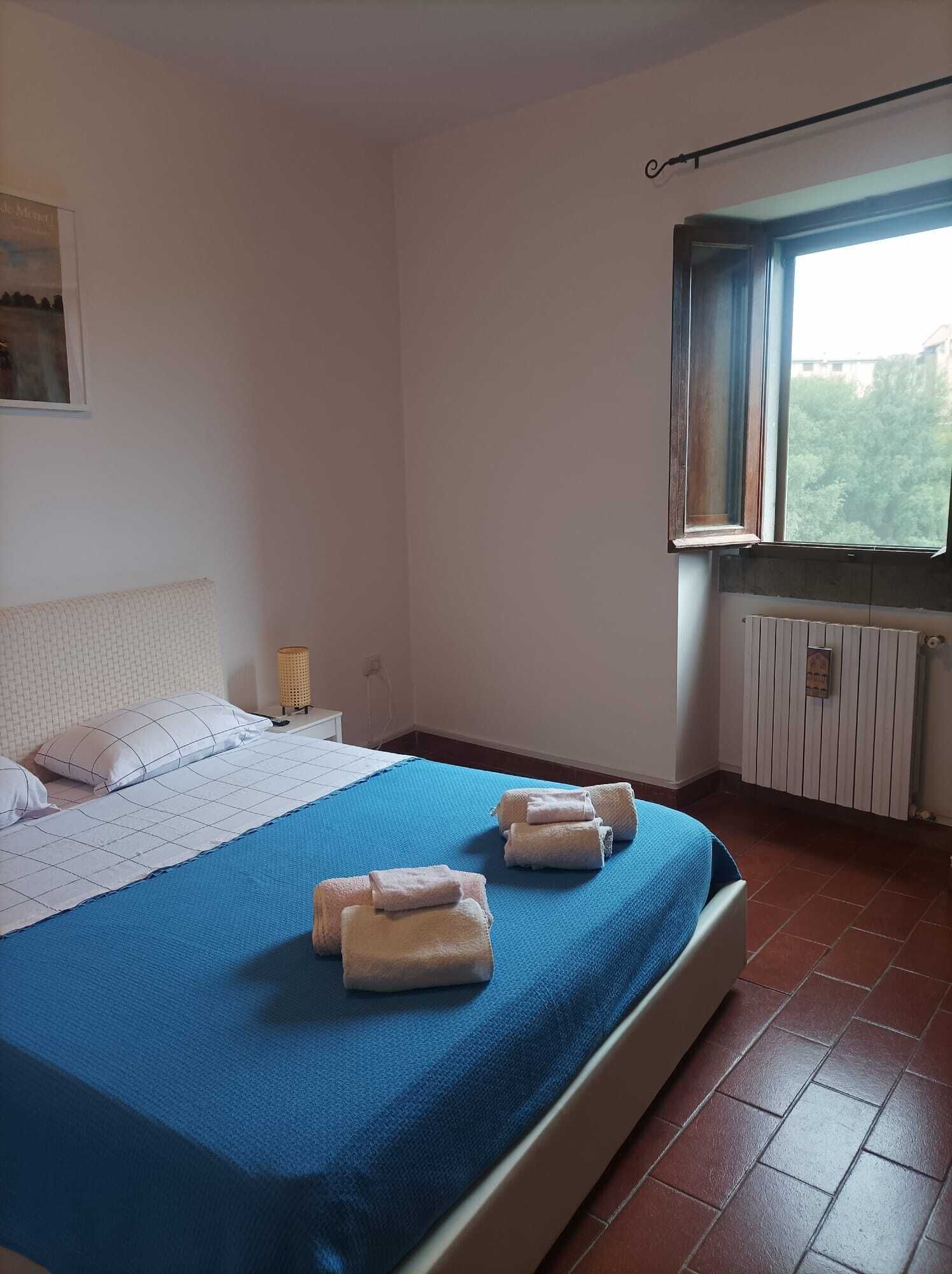 Ein Schlafzimmer mit einem Bett, Kissen, einem Nachttisch, einer Lampe und einem Fenster mit Vorhängen.
