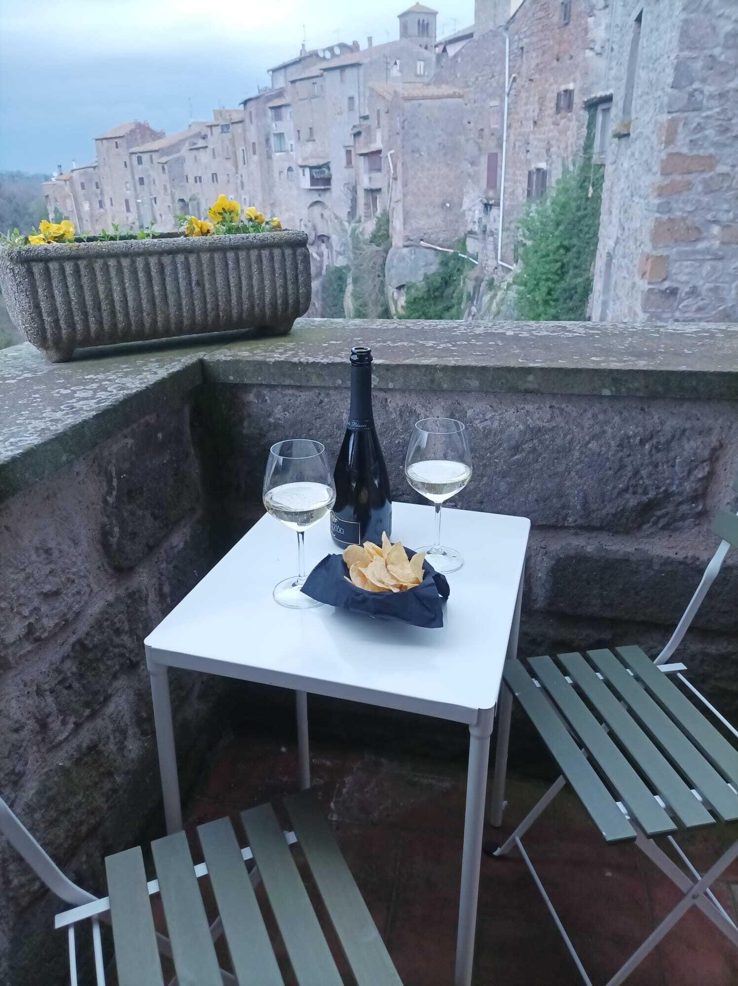A table with two wine glasses, a bottle, and a bowl of chips on a balcony overlooking a historic stone building.