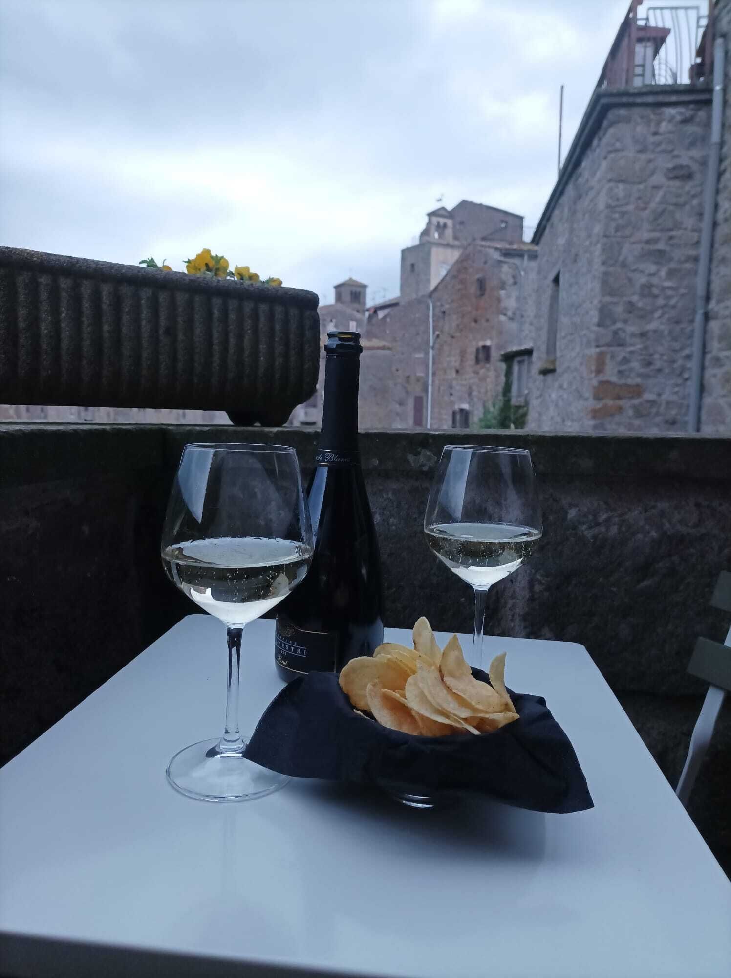 A table with two wine glasses and a bottle of wine, a basket of chips, and a view of old buildings in the background.