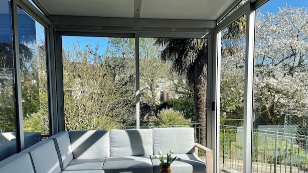A modern outdoor seating area with a sofa and a small table, surrounded by large windows offering a view of trees and a fence. - Charming 4-bedroom house with garden, close to shops and bus stop (Bruz)