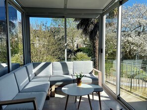 A modern outdoor seating area with a sofa and a small table, surrounded by large windows offering a view of trees and a fence. - Charming 4-bedroom house with garden, close to shops and bus stop (Bruz)