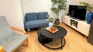 A living room with a blue sofa, a round black table with a book and vase, a TV on a white stand, and a potted plant. - Charming 4-bedroom house with garden, close to shops and bus stop (Bruz)
