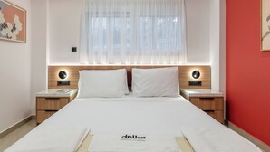 A neatly arranged hotel room with a bed, two bedside tables, a nightstand, a window with sheer curtains, and a red accent wall. - Glyfada Coastside Apartment (Glyfada)