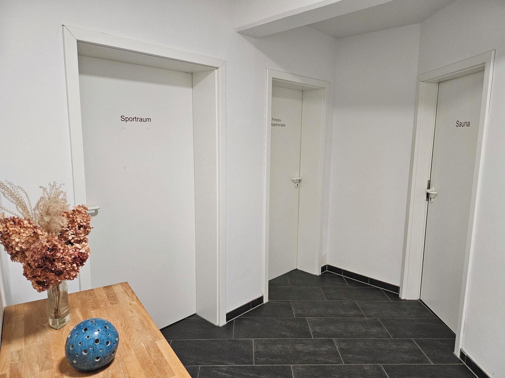 A hallway with three doors labeled "Spa," "Fitness," and "Spa." A wooden table with a vase of dried flowers and a blue ceramic bowl is visible.