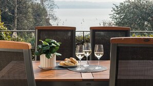 Uma mesa com uma planta em vaso, um prato de pastelaria e três copos de vinho, com o reflexo de um lago e árvores ao fundo.