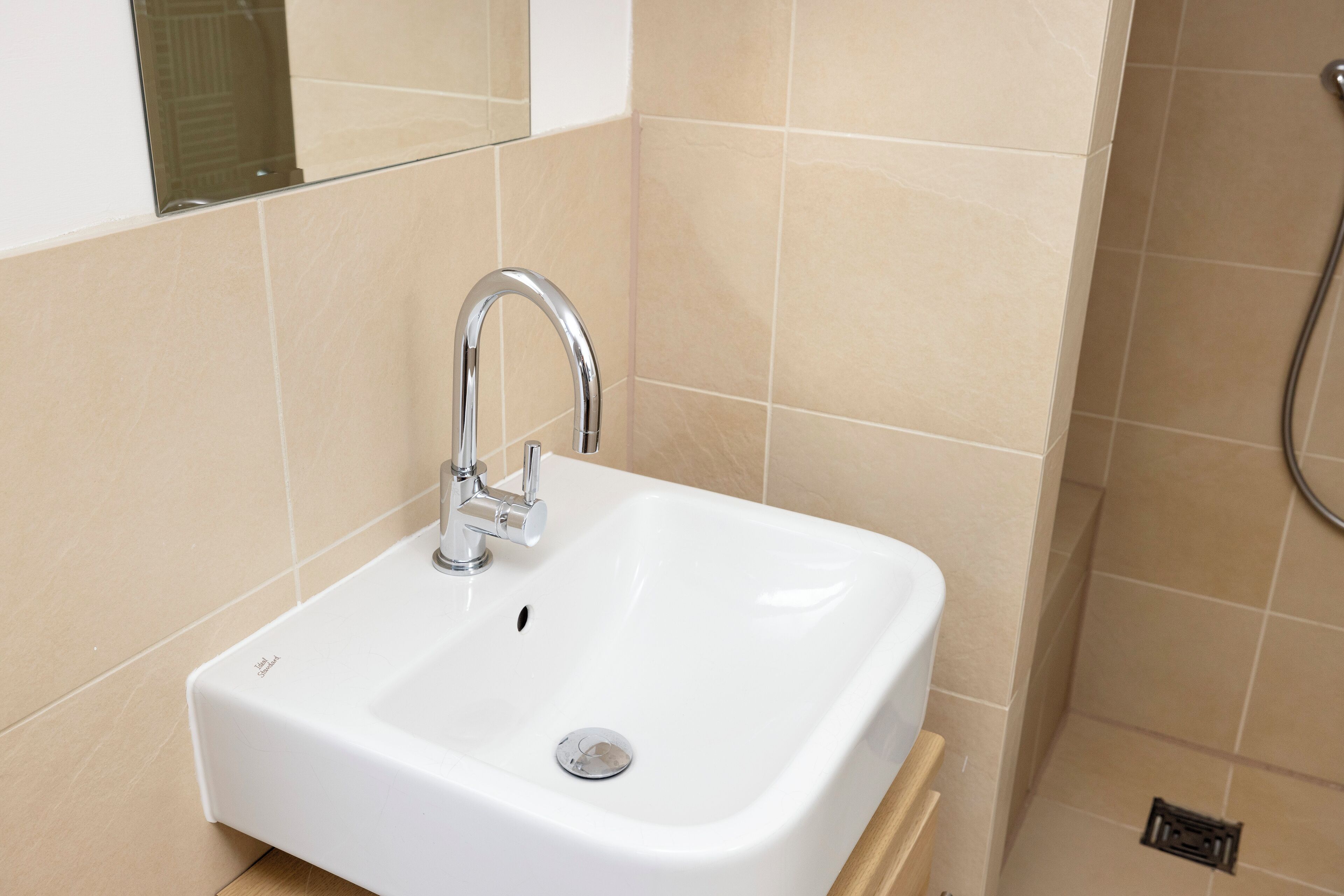 Un lavabo blanc dans la salle de bain, avec un robinet chromé, des murs recouverts de carreaux beiges et une zone douche située à droite.