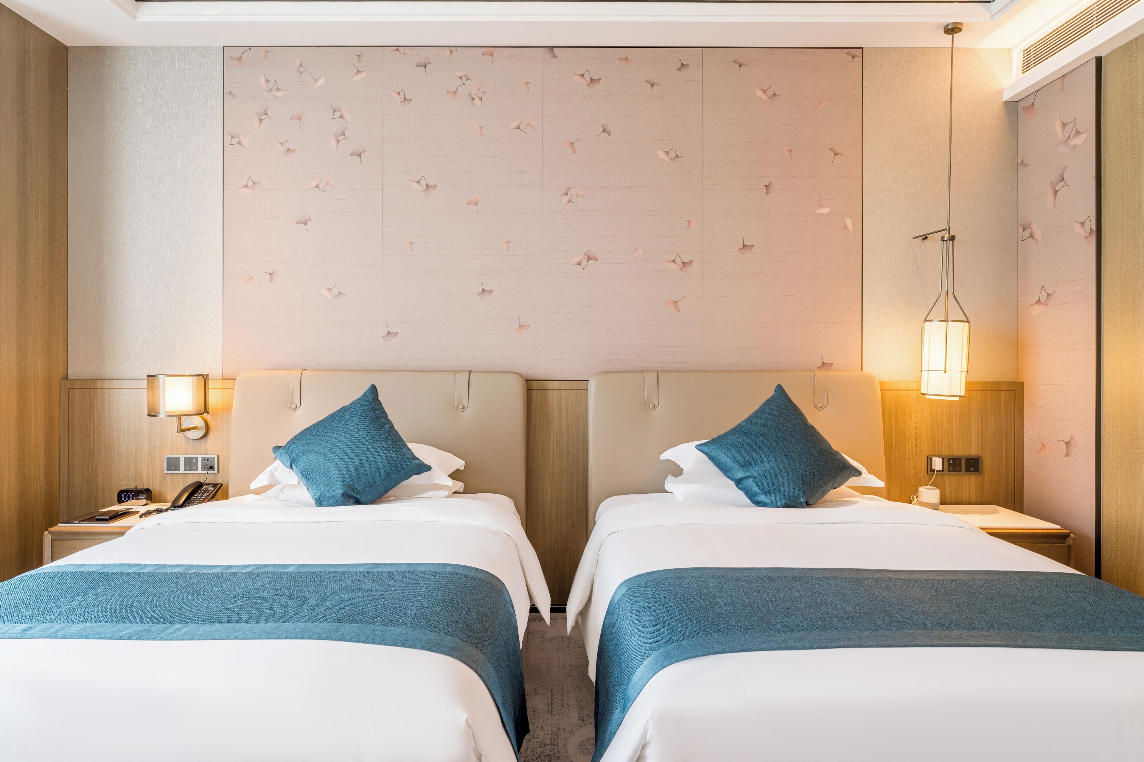 Een hotelkamer met twee bedden, een houten hoofdbord met een bloemendesign, bedlampjes, een telefoon op het nachtkastje en een klein tafeltje met een kopje en zakdoekje.