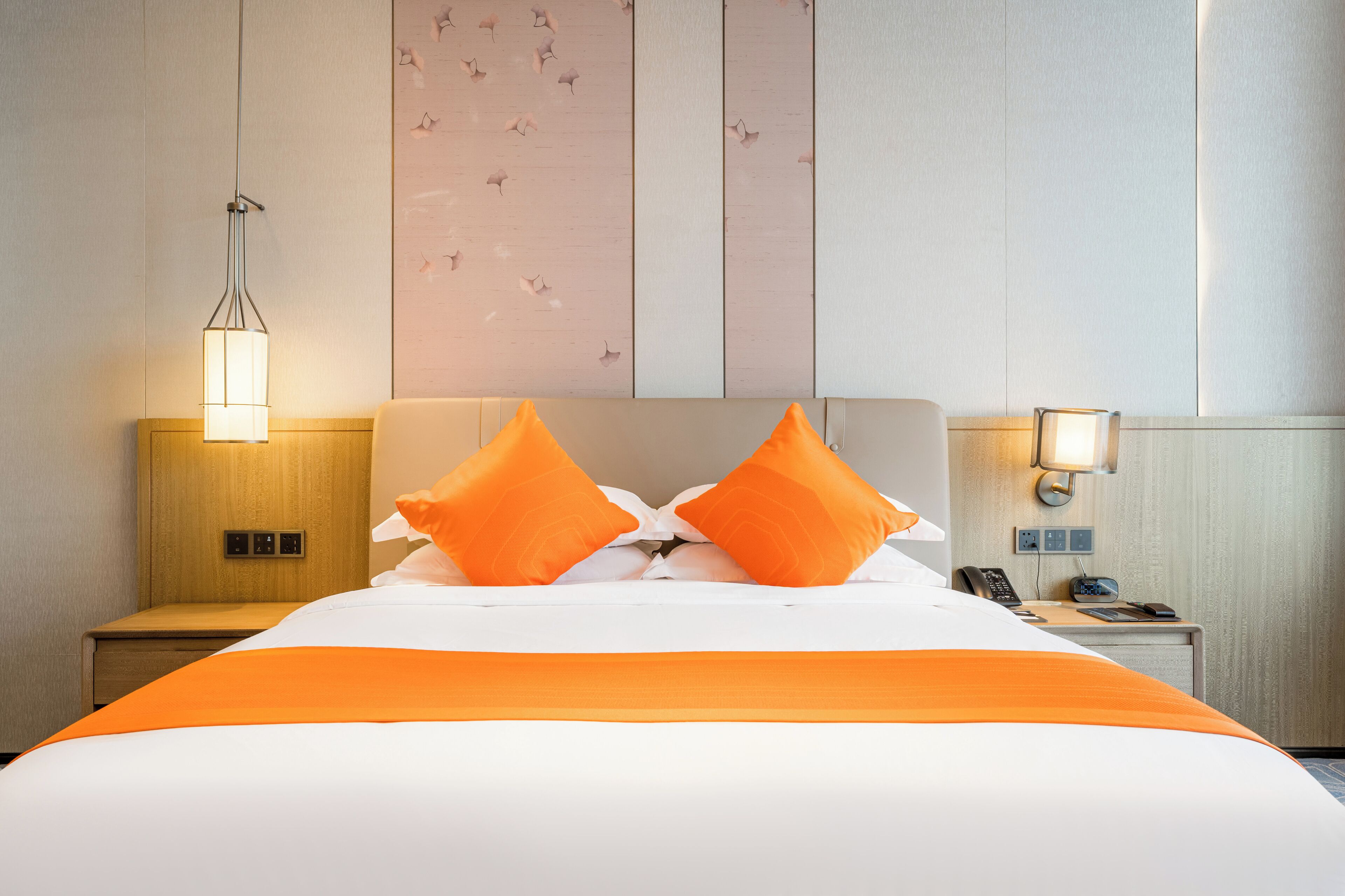 A hotel room with a bed, two orange pillows, a bedside lamp, a nightstand with a phone and a radio, and a wall with a floral pattern.