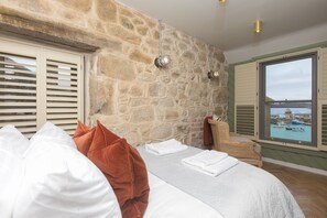A bedroom with a stone wall, a bed with white and brown pillows, a chair, and a view of boats through a window. - She Sells Sanctuary,St Ives–Sleeps 6–Sea View–Woodburner–Roll Top Steel Bath (St Ives)