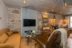A cozy living room with a stone wall, leather furniture, a flat-screen TV, and a fireplace. - She Sells Sanctuary,St Ives–Sleeps 6–Sea View–Woodburner–Roll Top Steel Bath (St Ives)