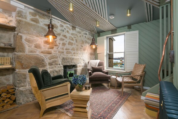 A cozy living room with a stone wall, wooden furniture, and a view through a window. - She Sells Sanctuary,St Ives–Sleeps 6–Sea View–Woodburner–Roll Top Steel Bath (St Ives)