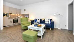 A modern living room with a sofa, armchair, and ottoman. There is a dining table with a bowl of fruit and a bottle. The room has wooden flooring and a kitchen in the background. - Comfortable vacation apartment with sea view and terrace in the Blue House in Ralswiek (Ralswiek)