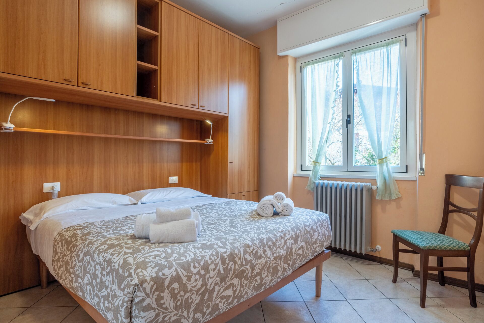 Un dormitorio con cama, una silla, una ventana con cortinas y armarios de madera.