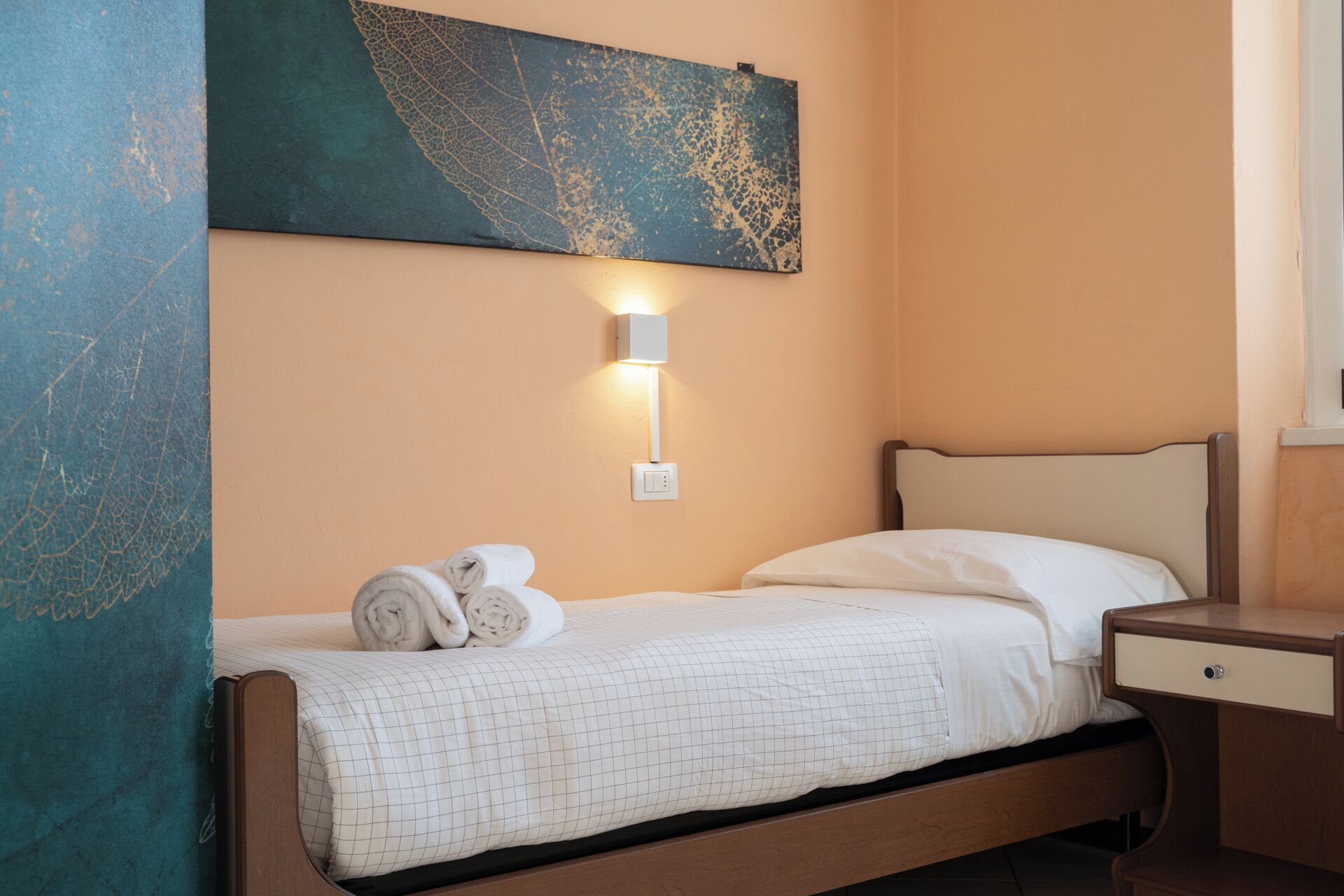 A neatly made bed with a white grid-patterned comforter and rolled towels, a wall-mounted light fixture, and a large abstract painting above the bed.