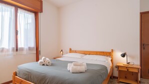 Un lit bien fait, avec deux serviettes enroulées, une lampe de chevet et une table de nuit en bois, dans une pièce dotée d’une fenêtre et d’une porte.