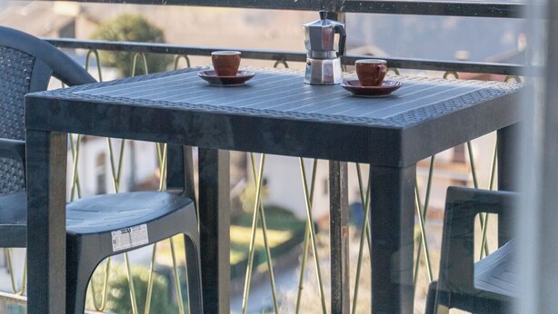 Uma varanda com mesa e cadeiras, uma cafeteira e xícaras. Uma paisagem montanhosa é visível ao fundo.