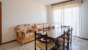Uma área de refeições com uma mesa redonda e quatro cadeiras de madeira, dois sofás florais e uma janela grande com cortinas leves.
