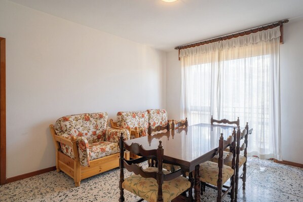 A dining area with a round table and four wooden chairs, two floral sofas, and a large window with sheer curtains. - Residence Bellavista - Stambecco (P2) (Barzio)