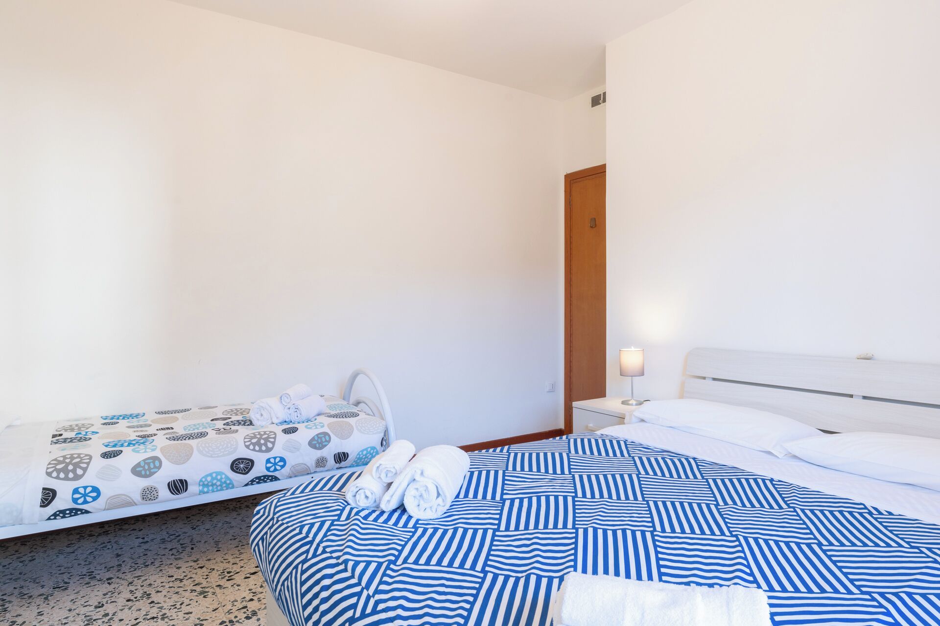 Une chambre d’hôtel avec deux lits, l’un recouvert d’une couvre-lit à motifs et l’autre d’une couvre-lit à rayures. On y trouve une table de chevet avec une lampe et une porte en bois.