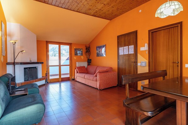 A spacious living area with a fireplace, a sofa, a dining table, and a view through a large sliding door. - Spacious mountain view attic apartment (Barzio)
