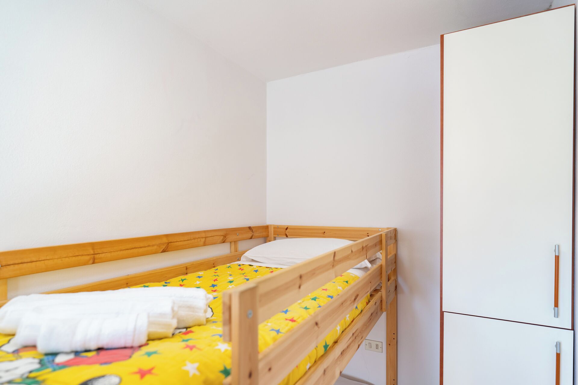 A small, single-bed room with a wooden bunk bed, a white wardrobe, and a colorful bedspread.