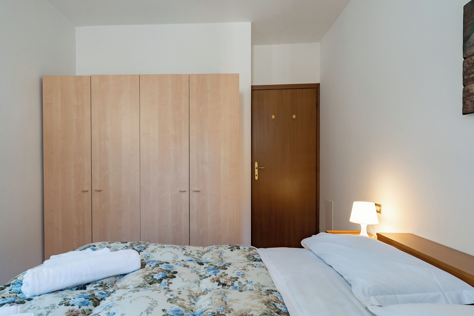 A hotel room with a bed, a wooden wardrobe, and a nightstand with a lamp.