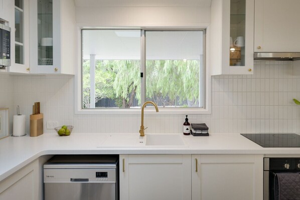 A modern kitchen with white cabinetry, a stainless steel dishwasher, a double sink with a gold faucet, and a window overlooking greenery. - The House - Dunsborough (Dunsborough)