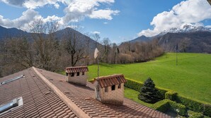 A view of a mountainous landscape with a clear sky, a green field, and a building with a tiled roof and two small brick structures. - Mountain lodging with mountain view (Barzio)