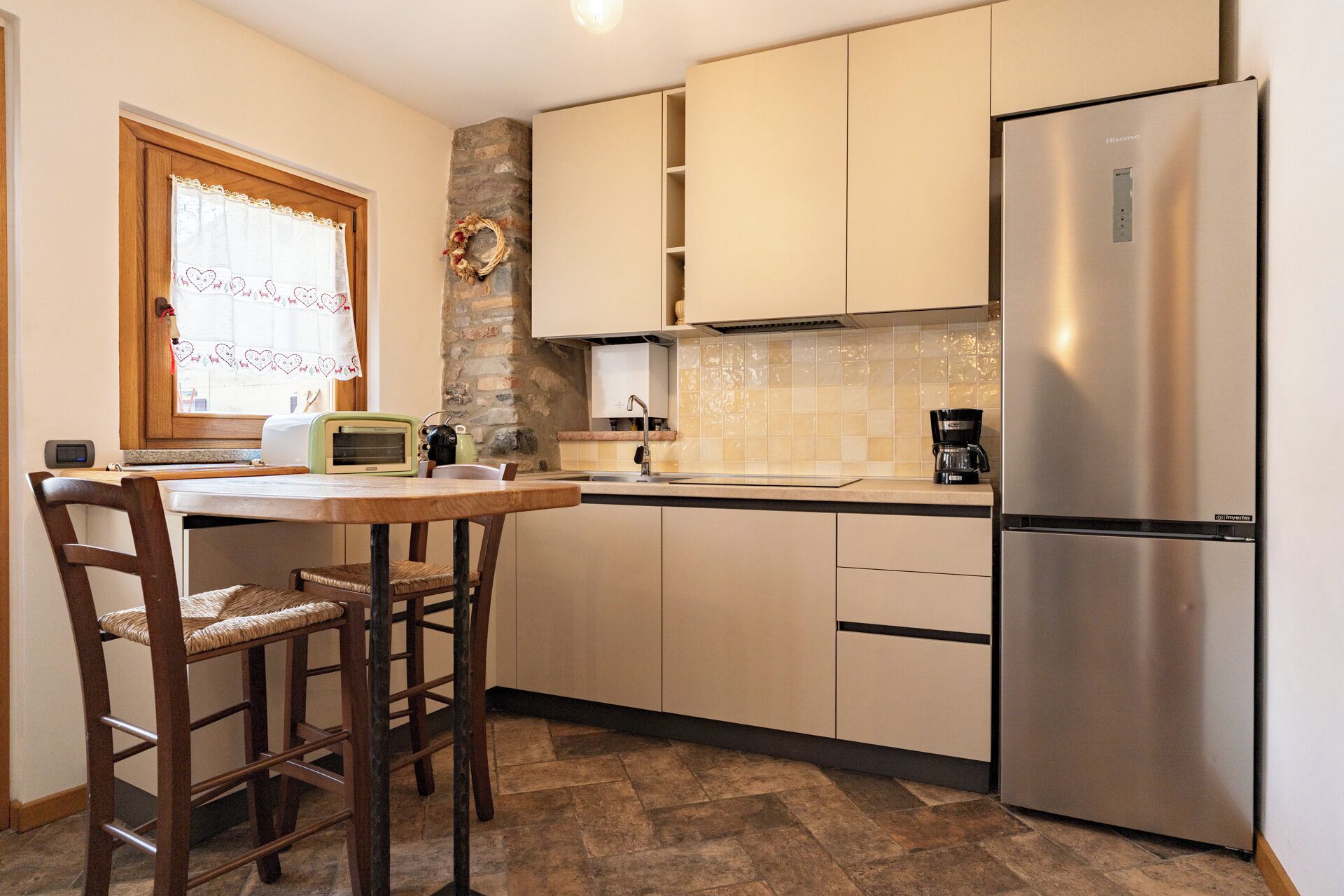 Une cuisine compacte dotée d’un réfrigérateur intégré, d’un mur décoratif en pierre et d’une fenêtre agrémentée d’un rideau à motifs en forme de cœur.