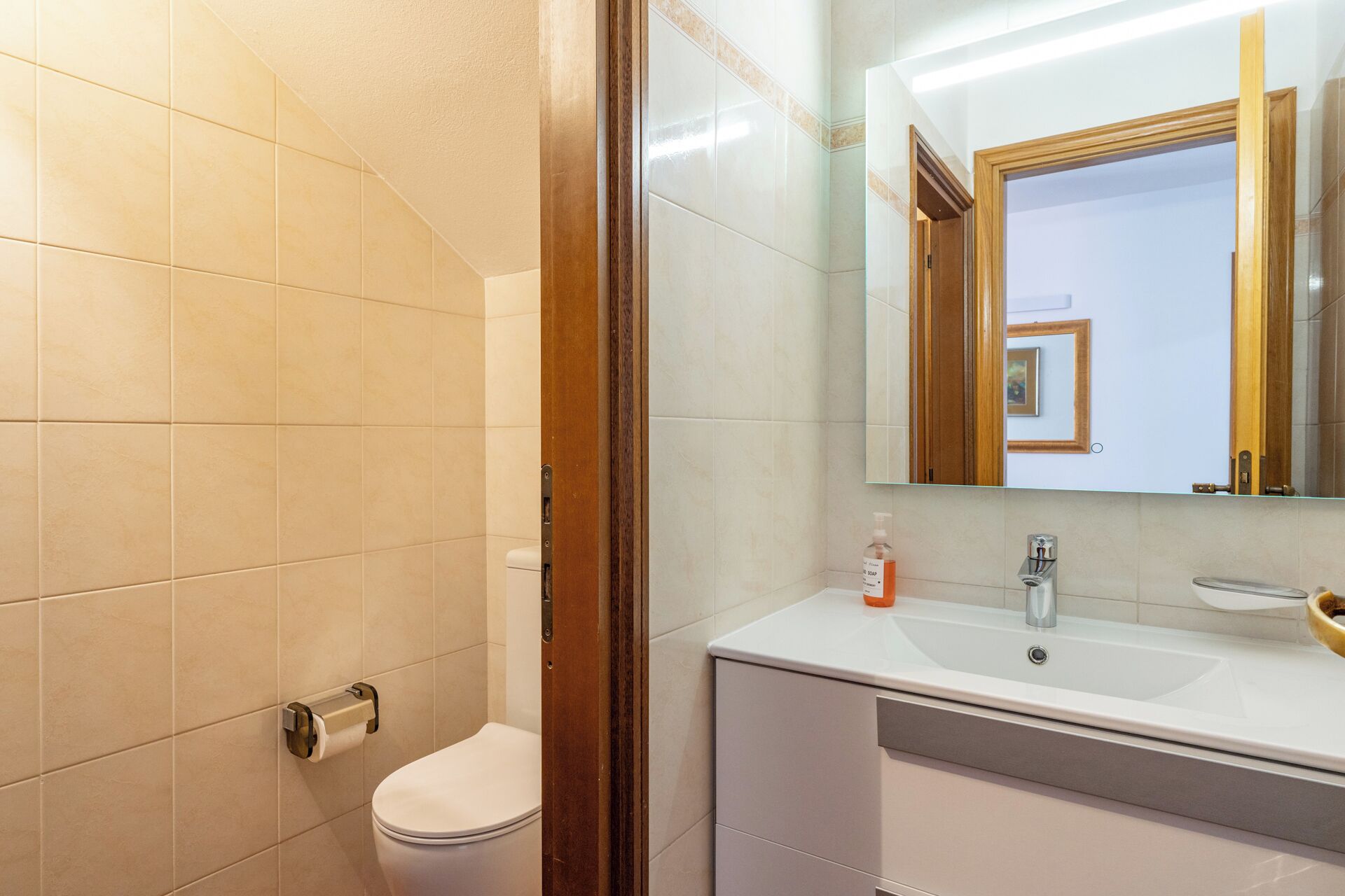 A bathroom with beige tiles, a white toilet, a sink with a mirror, and a bottle of soap.
