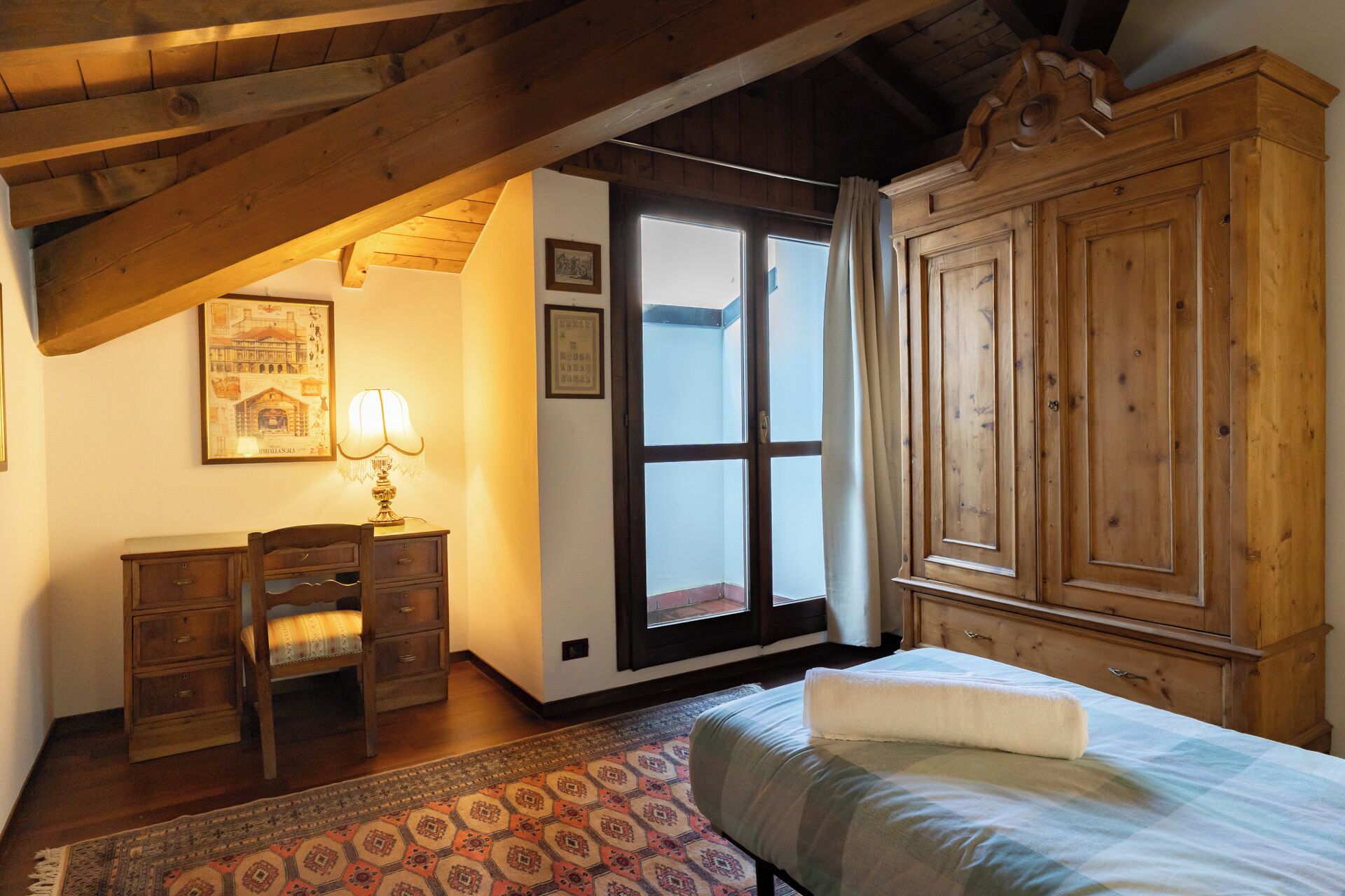 Una camera da letto con un letto, un armadio in legno, una scrivania con una sedia, una lampada e un tappeto decorato.